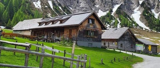 Bergiges Gebiet mit einem Holzhaus, einem Zaun und schneebedeckten Gipfeln. Das Haus hat ein geneigtes Dach und einen Schornstein.