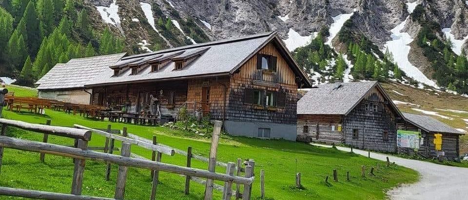 Bergiges Gebiet mit einem Holzhaus, einem Zaun und schneebedeckten Gipfeln. Das Haus hat ein geneigtes Dach und einen Schornstein.