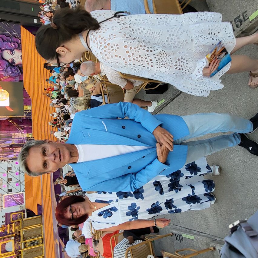 A man in a blue suit stands with his hands on his hips, smiling at the camera. Behind him, a woman in a white dress leans over to look at him. They are in a crowded room with people sitting in chairs.