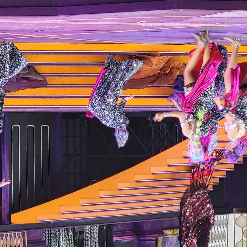 A group of performers in sequined costumes are performing acrobatics on a stage with a yellow and purple backdrop.