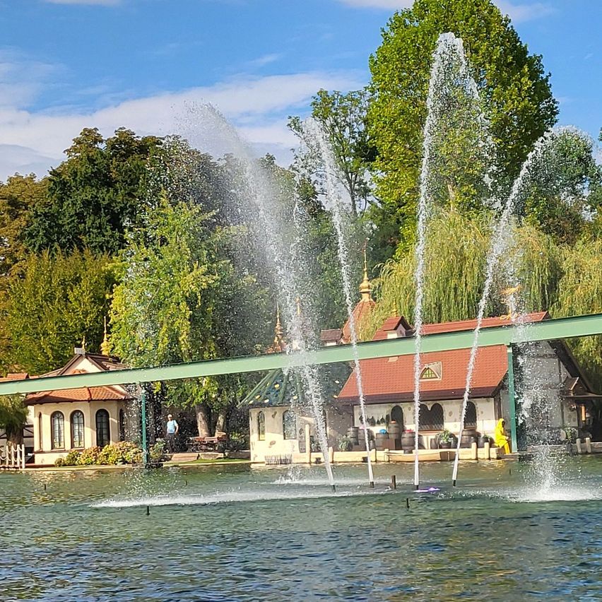 Ein Wasserbrunnen in einem Park mit einer Brücke und Gebäuden im Hintergrund. Bäume und ein blauer Himmel sind sichtbar.