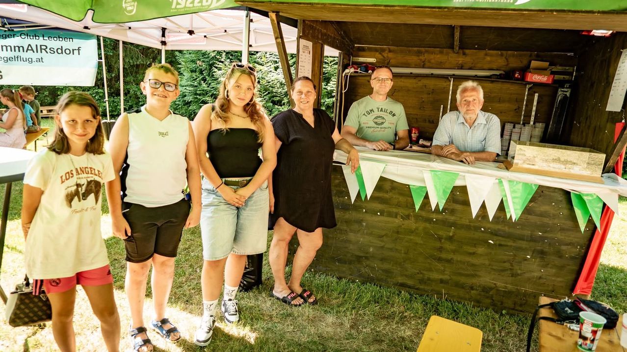 Five individuals pose for a picture in front of a wooden booth with green bunting and a white canopy, under a clear blue sky.