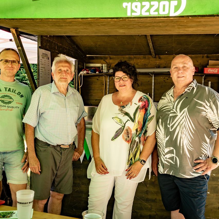 Four people stand outside a small wooden shop. A woman in white, a man in green, a man in glasses, and a man in a floral shirt. They appear to be posing for a picture.