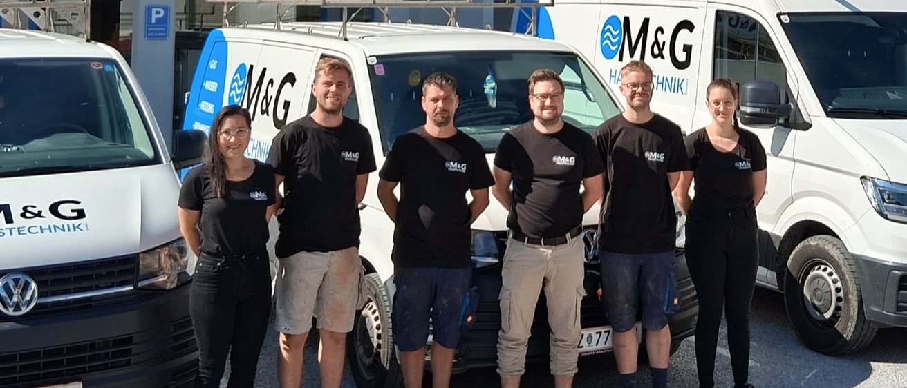 Eine Gruppe von Menschen in schwarzer Kleidung posiert vor drei Vans, zwei davon mit dem M&G Haustechnik-Logo. Die Vans sind in einer Reihe vor einem Gebäude mit weißen Fenstern und einem Schild mit der Aufschrift M&G geparkt.