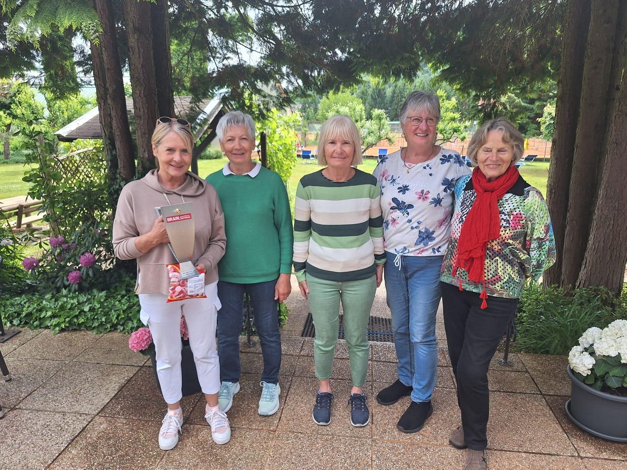 Fünf Frauen stehen zusammen in einem Garten, lächeln und posieren für ein Foto. Sie tragen Freizeitkleidung und eine hält einen Pokal. Hinter ihnen stehen Bäume, Pflanzen und ein Gebäude.