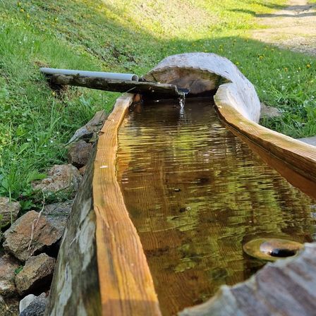 Ein rustikales hölzerne Wasserbecken auf einer Grasfläche mit Steinen, gefüllt mit klarem Wasser, mit einem Rohr, das dazuführt.