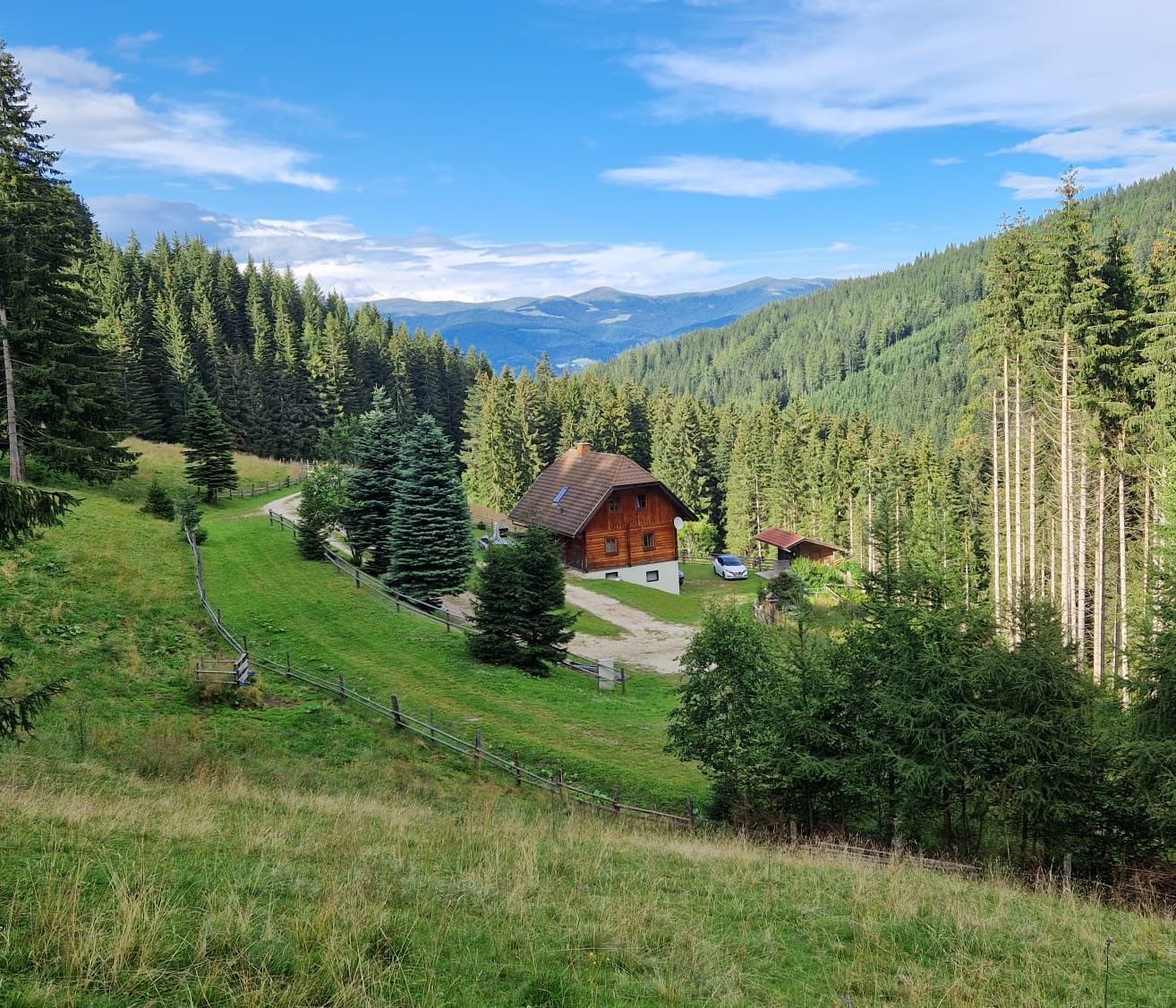 Eine kleine Blockhütte umgeben von grünen Bäumen und Gras. Ein Auto steht vor der Hütte. In der Ferne gibt es Berge.