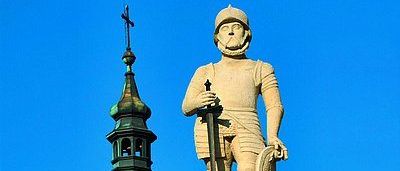 Eine Statue eines Ritters steht vor einem Glockenturm mit einem grünen Turm. Der Ritter hält ein Schwert und einen Schild. Der Turm hat eine Uhr und einen Glockenturm. Bäume und Gebäude umgeben das Gebiet.