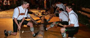 Mehrere Männer in traditioneller bayerischer Kleidung sägen einen Baumstamm, während sie Hämmer halten, und eine Band spielt im Hintergrund.
