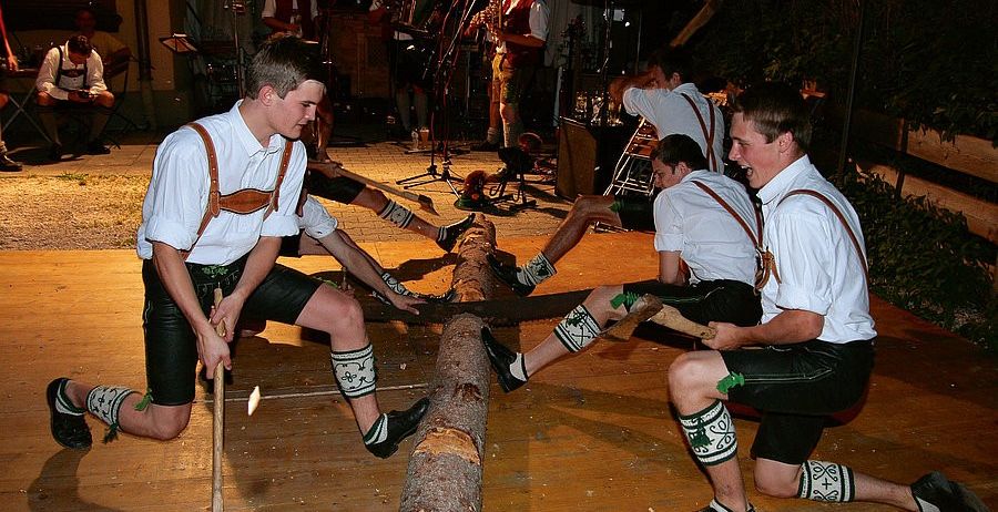 Mehrere Männer in traditioneller bayerischer Kleidung sägen einen Baumstamm, während sie Hämmer halten, und eine Band spielt im Hintergrund.