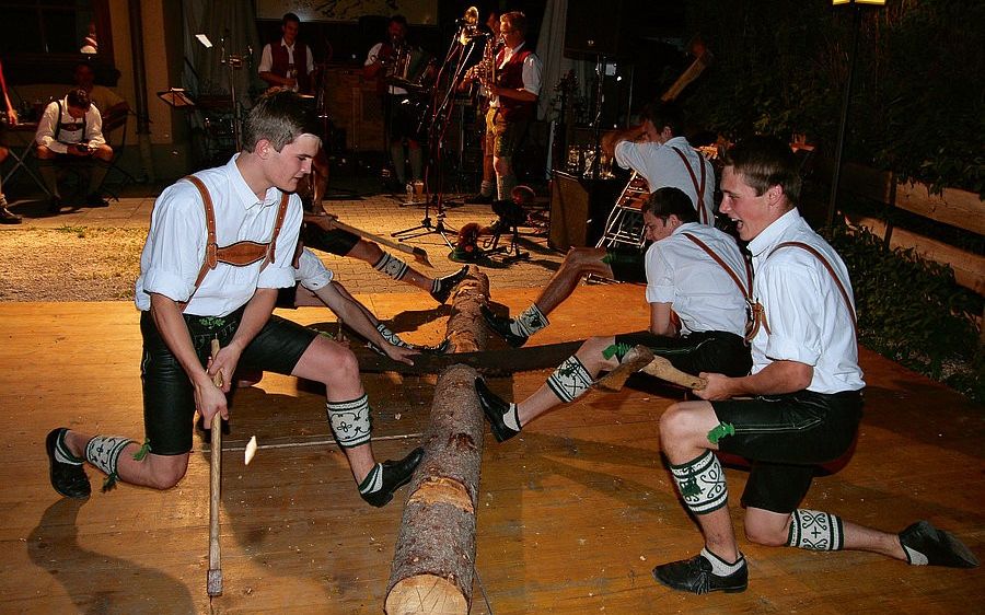 Mehrere Männer in traditioneller bayerischer Kleidung sägen einen Baumstamm, während sie Hämmer halten, und eine Band spielt im Hintergrund.