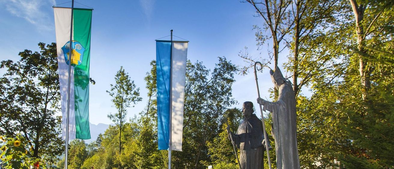 Zwei Statuen von Heiligen, eine mit einem Stab, stehen auf Felsen neben einem Brunnen. Zwei Fahnen, eine grün und weiß, und eine blau und weiß, stehen in der Nähe. Die Szenerie ist vor einem Hintergrund aus üppigen Bäumen und einem klaren blauen Himmel.