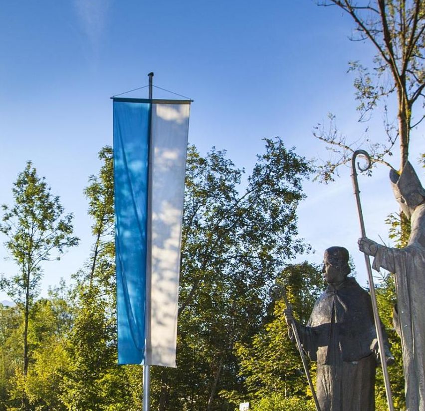 Zwei Statuen von Heiligen, eine mit einem Stab, stehen auf Felsen neben einem Brunnen. Zwei Fahnen, eine grün und weiß, und eine blau und weiß, stehen in der Nähe. Die Szenerie ist vor einem Hintergrund aus üppigen Bäumen und einem klaren blauen Himmel.