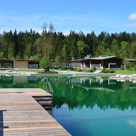 Ein ruhiger Pool mit einem Holzsteg und einem modernen Gebäude, umgeben von üppigem Grün.