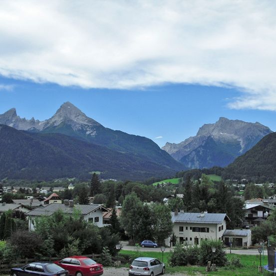 Ein Bergdorf mit mehreren Häusern und geparkten Autos davor, umgeben von Bäumen und Bergen unter einem teilweise bewölkten Himmel.