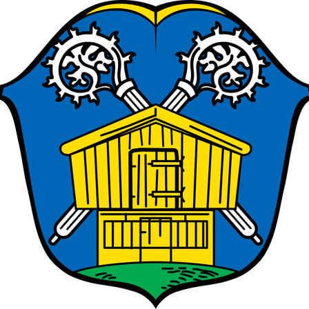 Das Wappen zeigt ein gelbes Holzhaus mit einer Tür und Fenstern auf blauem Hintergrund. Oben auf dem Haus befinden sich zwei weiße Zahnräder. Das Haus ist von grünem Gras umgeben.