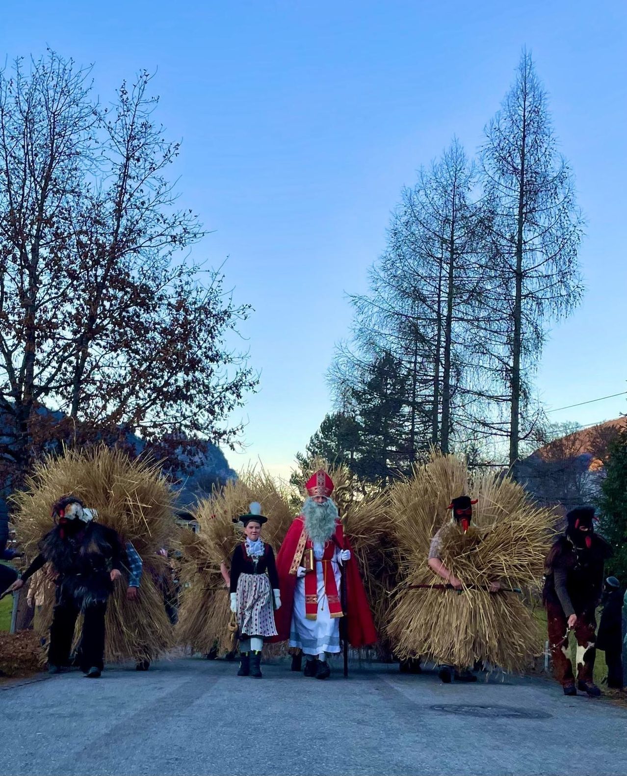 Eine Gruppe von Menschen in Kostümen, einige mit Masken, geht auf einer Straße. Sie tragen große Bündel Stroh. Dahinter säumen kahle Bäume die Straße. In der Ferne sind Berge zu sehen.