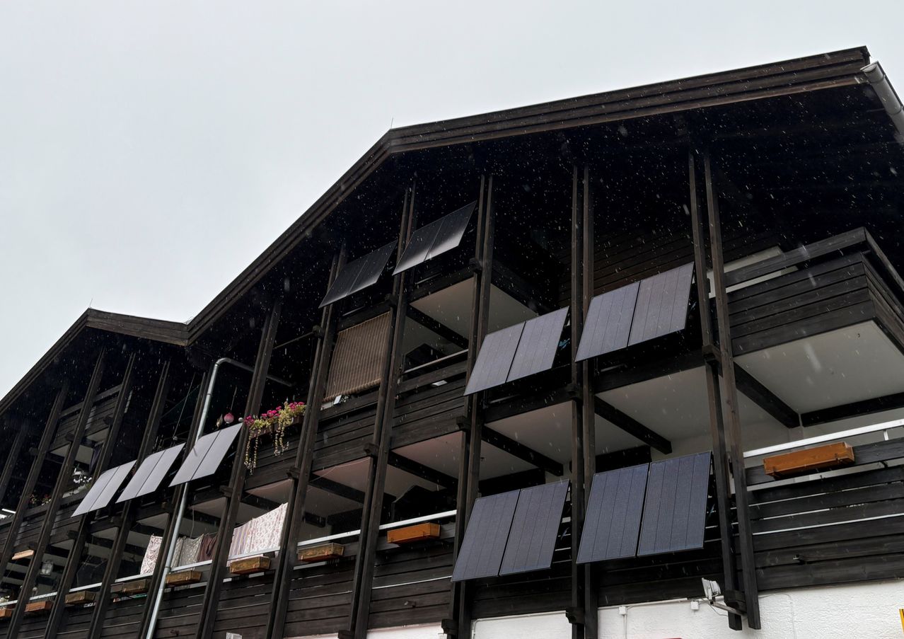 Ein modernes Gebäude mit einem dreieckigen Dach hat mehrere Solarmodule an seinen Seiten. Es schneit, und die Module sind leicht geneigt. Jede Etage hat einen Balkon mit einer Brüstung, und einige Balkone haben Blumenkästen.