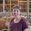 Eine Frau in einer Scheune hält einen Holzstab und lächelt, während sie neben Kühen steht, von denen einige Heu fressen. Dahinter stützen Holzbalken und Metallpfosten die Struktur.