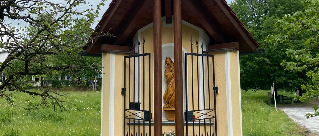 Eine kleine Kapelle mit einer Statue der Jungfrau Maria und einem Wetterhahn auf dem Dach. Sie hat ein schwarzes Tor und ist von Gras und Bäumen umgeben.