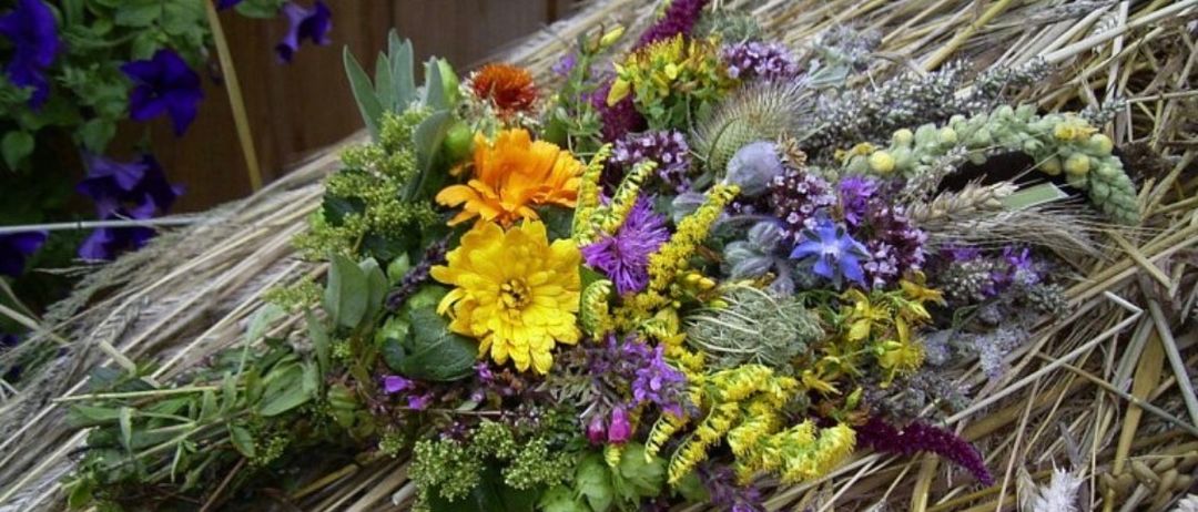 Ein farbenfroher Strauß aus Wildblumen und Kräutern liegt auf einem Strohbett, wobei lila und gelbe Blumen im Arrangement hervorstechen.