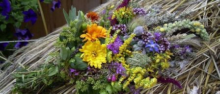 Ein farbenfroher Strauß aus Wildblumen und Kräutern liegt auf einem Strohbett, wobei lila und gelbe Blumen im Arrangement hervorstechen.