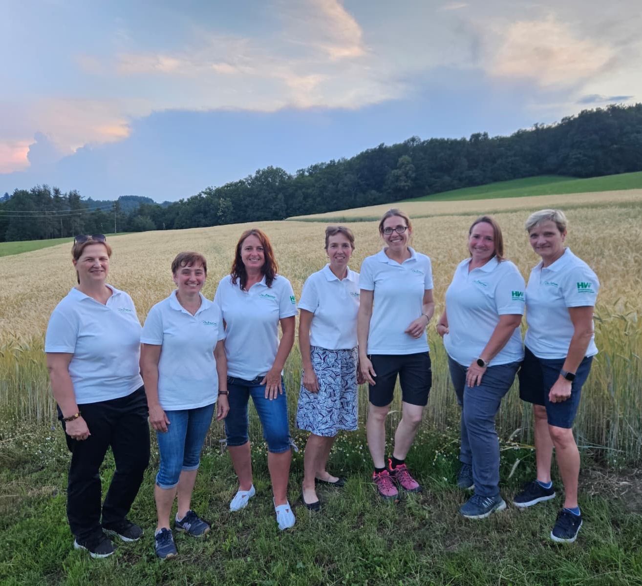 Sieben Frauen in weißen Shirts stehen in einem Feld mit einem Sonnenuntergang im Hintergrund. Sie lächeln und scheinen für ein Foto zu posieren.