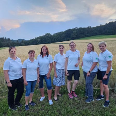 Sieben Frauen in weißen Shirts stehen in einem Feld mit einem Sonnenuntergang im Hintergrund. Sie lächeln und scheinen für ein Foto zu posieren.