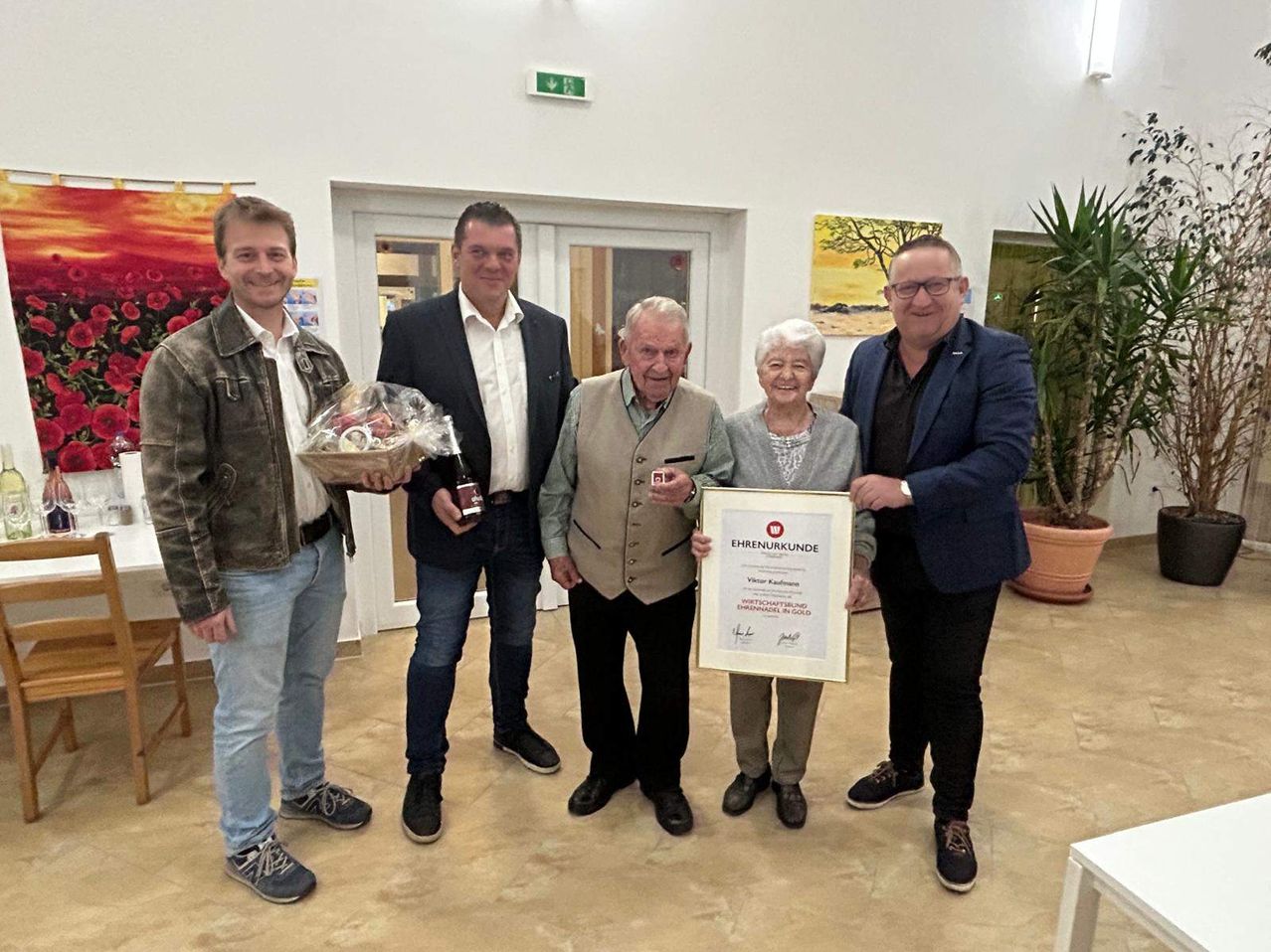 Fünf Menschen stehen lächelnd zusammen in einem Raum und posieren für ein Foto. Ein älteres Paar hält ein Zertifikat, während andere Geschenke halten. Eine Pflanze und ein gerahmtes Bild sind im Hintergrund.