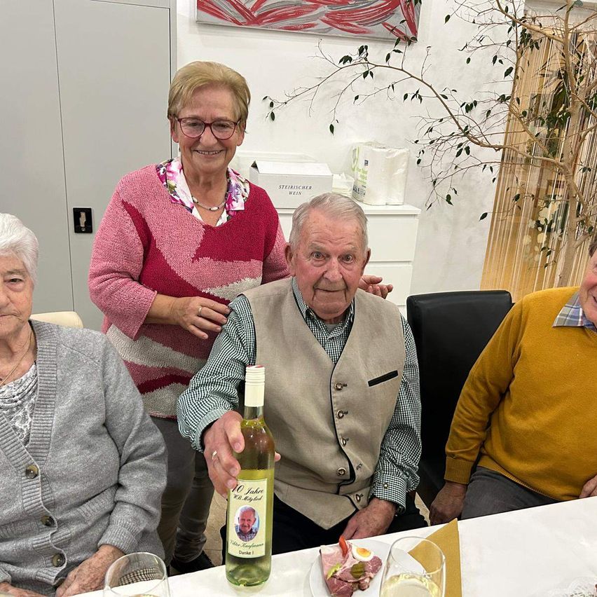 Drei ältere Personen sitzen an einem Tisch und feiern einen Jahrestag. Der Mann hält eine Flasche Wein, und es gibt Teller mit Essen und Gläser auf dem Tisch. Hinter ihnen ist eine weiße Wand mit einem Bilderrahmen und einer Pflanze.