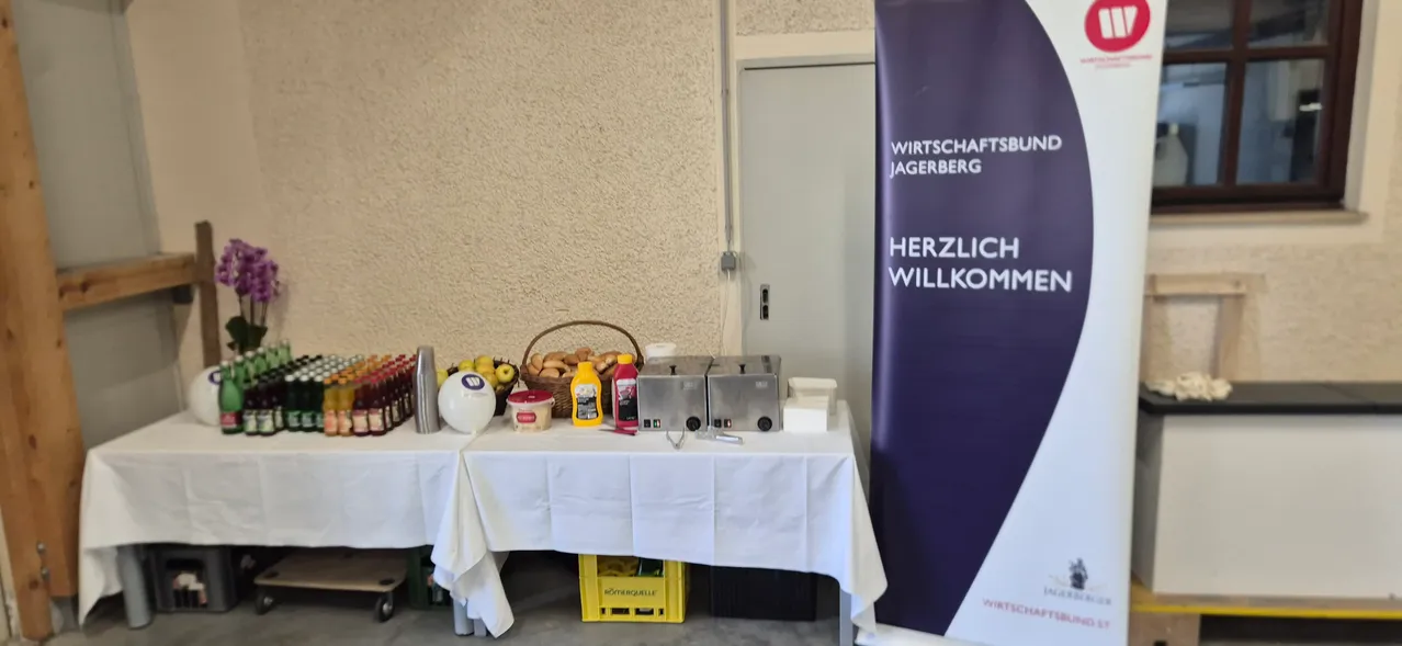 Ein Tisch mit weißer Tischdecke, mit Lebensmitteln und Getränken, in der Nähe eines Wirtschaftsberg-Banners.