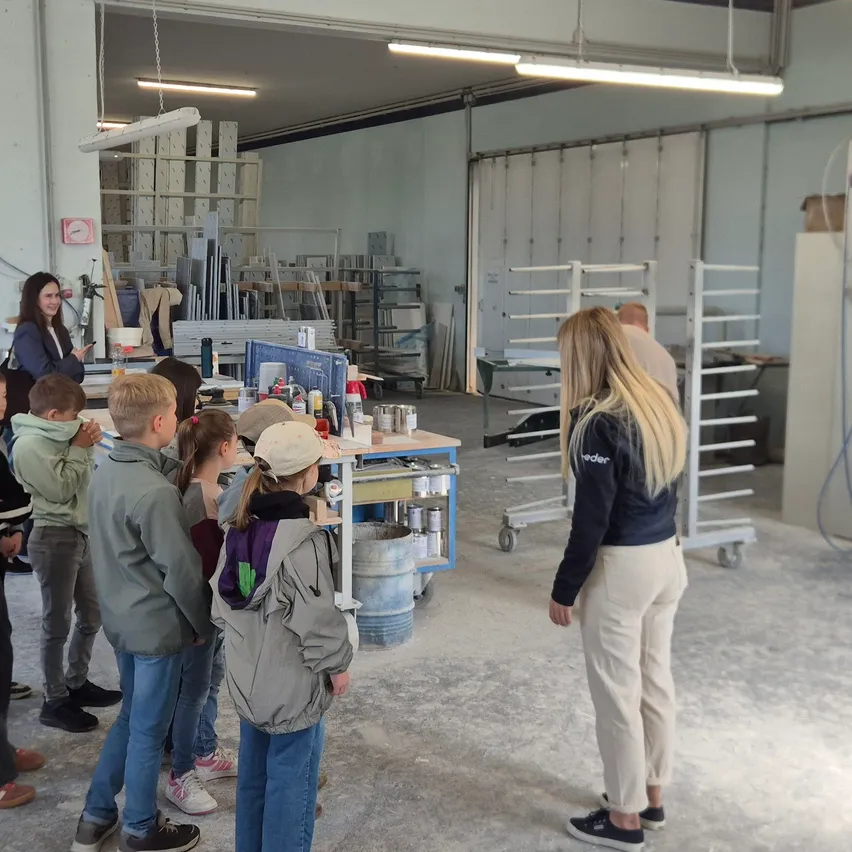 In einer Werkstatt stehen eine Gruppe von Kindern und Erwachsenen um einen Tisch mit verschiedenen Gegenständen herum und hören einer Frau zu, die spricht. Im Hintergrund sind Geräte und Regale zu sehen.
