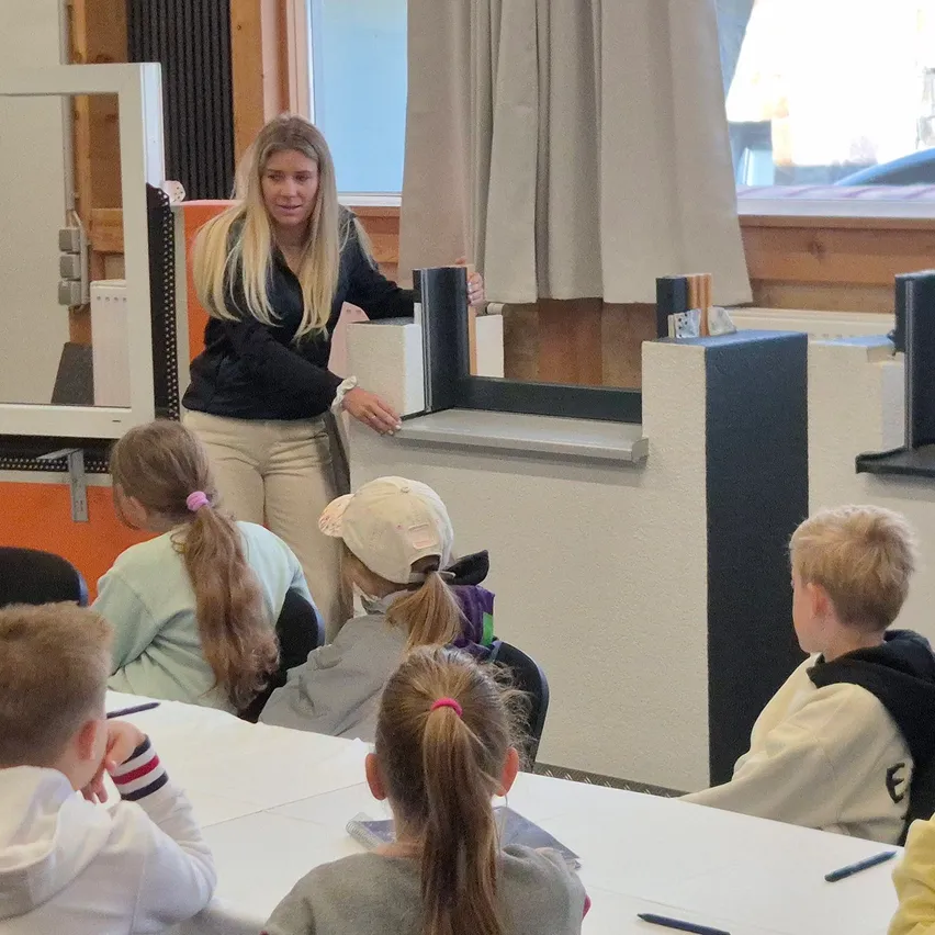 Eine Frau steht hinter einem Schreibtisch und spricht eine Gruppe von Kindern an, die an einem Tisch sitzen, wahrscheinlich in einem Klassenzimmer.