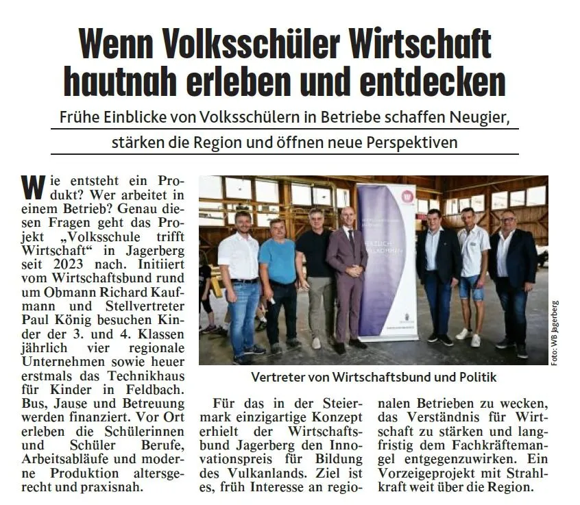 Eine Gruppe von Menschen, einschließlich eines Mannes im Anzug, steht vor einem Banner und einem Gebäude. Das Banner hat Text auf Deutsch. Der Mann im Anzug schaut in die Kamera. Das Bild bezieht sich auf ein Projekt namens 'Volksschule trifft Wirtschaft'.