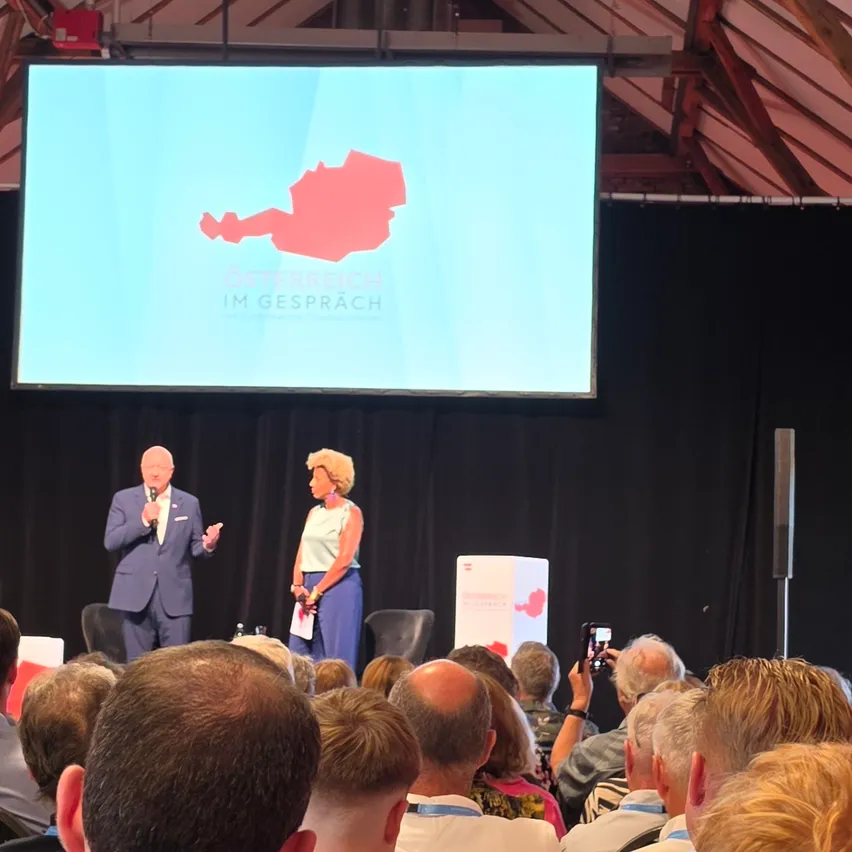Ein Mann und eine Frau stehen auf einer Bühne mit einer großen Leinwand dahinter und präsentieren einem Publikum. Die Leinwand zeigt eine Karte von Österreich mit dem Text 'Österreich im Gespräch'.