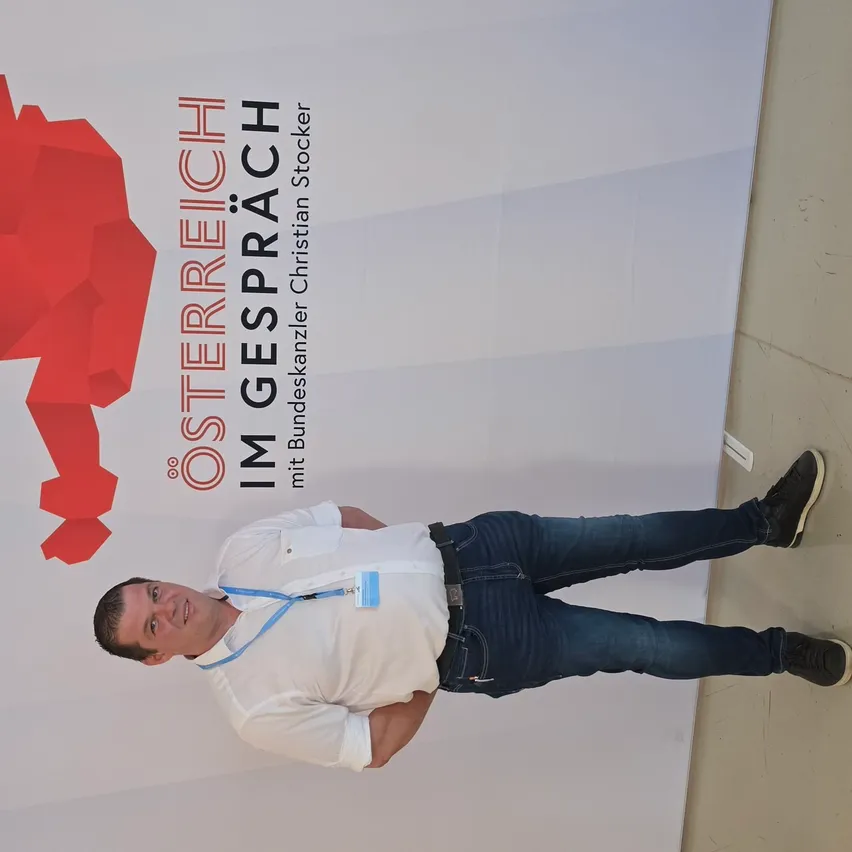 Ein Mann in einem weißen Hemd und blauen Jeans steht vor einem weißen Hintergrund mit einem roten geometrischen Design und Text auf Deutsch.
