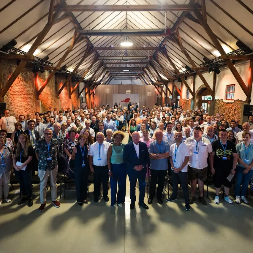 Eine große Gruppe von Menschen in formeller und lässiger Kleidung steht in einem großen Raum mit Holzbalken und Ziegelwänden, möglicherweise für eine Konferenz.