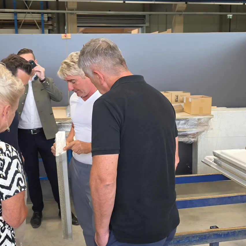 Eine Gruppe von Menschen in einer Fabrikumgebung, ein Mann in einem schwarzen T-Shirt inspiziert ein Material, während andere zusehen.