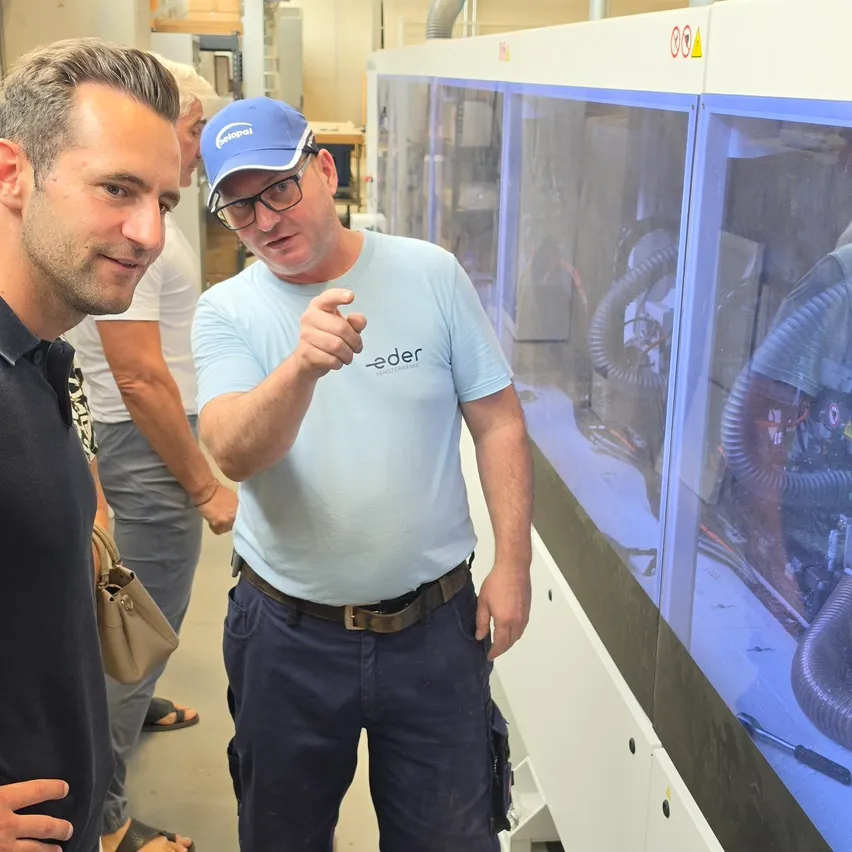 Zwei Männer in einer Fabrikumgebung, einer trägt ein blaues Shirt mit dem Wort 'eder' und eine blaue Kappe und zeigt auf eine große Maschine mit Glasfront.