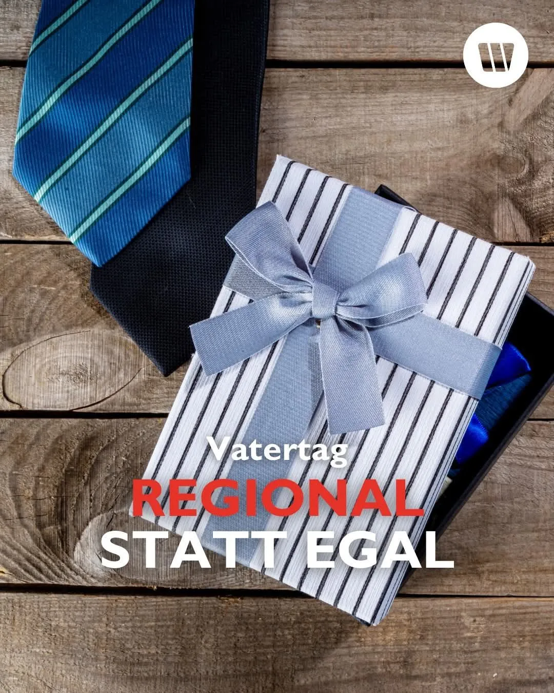 Eine Geschenkbox mit einem blauen Band und gestreiftem Stoff liegt auf einer Holzoberfläche. Ein blauer Krawatte und eine schwarze Krawatte sind daneben. Der Text lautet 'Vatetag Regional Statt Egal'.