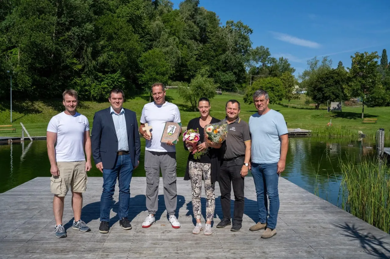 Sechs Menschen stehen auf einer Dock nahe einem See und lächeln. Eine Frau hält Blumen und einen Pokal. Der Mann neben ihr hält ein Buch. Der Mann in der Mitte trägt ein weißes T-Shirt. Die Männer an den Enden tragen Anzüge.