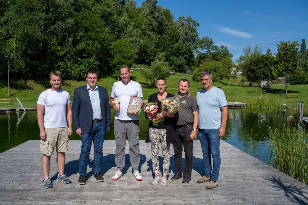 Sechs Menschen stehen auf einer Dock nahe einem See und lächeln. Eine Frau hält Blumen und einen Pokal. Der Mann neben ihr hält ein Buch. Der Mann in der Mitte trägt ein weißes T-Shirt. Die Männer an den Enden tragen Anzüge.