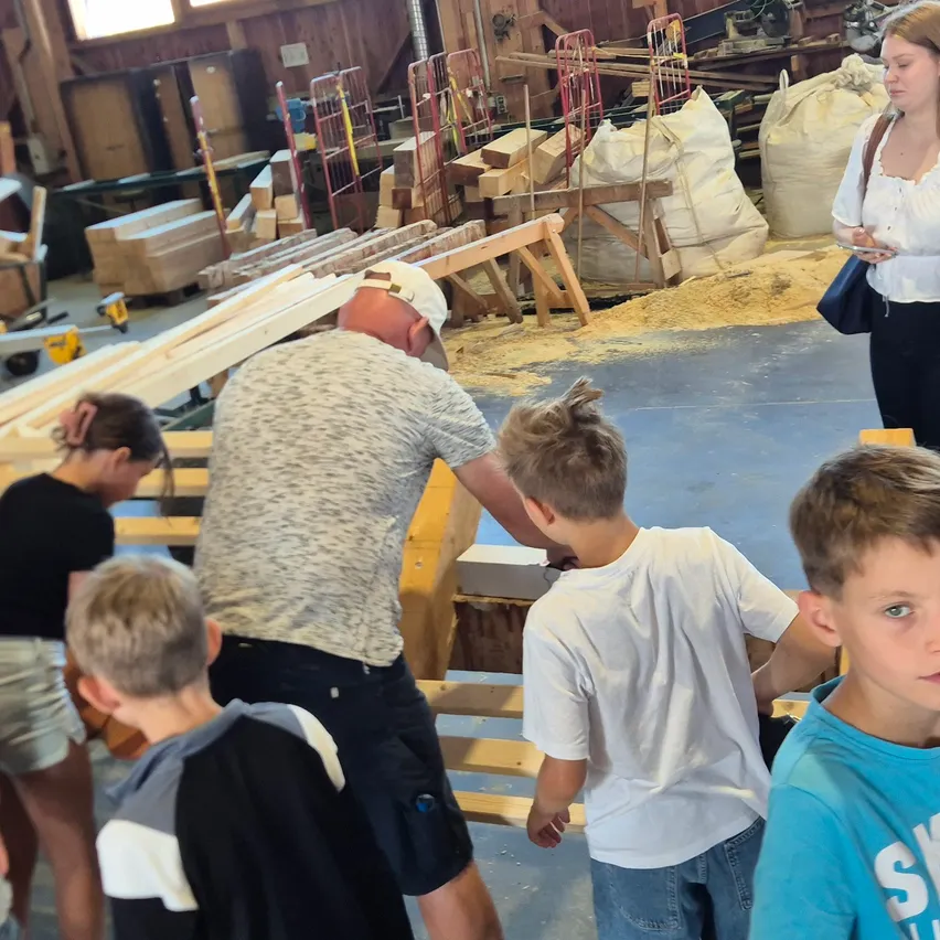 Eine Gruppe von Kindern und Erwachsenen befindet sich in einer Werkstatt. Ein Mann und eine Frau stehen, während andere eine Holzstruktur betrachten.