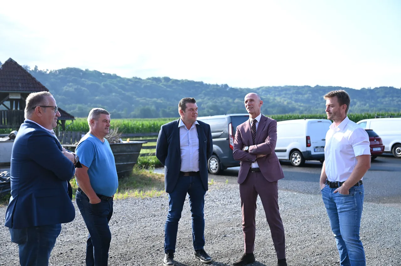 Fünf Männer stehen draußen, zwei in Anzügen. Einer hat die Arme verschränkt. Dahinter ist ein Lieferwagen geparkt, und ein LKW ist zu sehen. Im Hintergrund zeigt sich ein Feld und ein Berg.