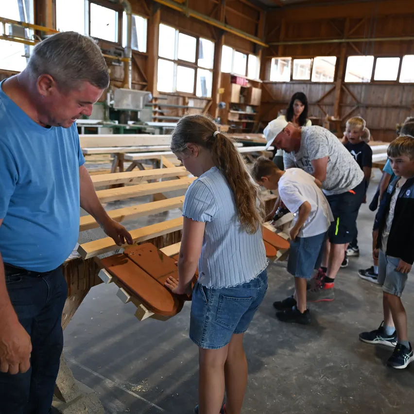 In einer Werkstatt arbeiten Erwachsene und Kinder an Holzstücken. Ein Mann und ein Mädchen konzentrieren sich auf einen Holzgegenstand. In der Nähe arbeiten andere an verschiedenen Holzgegenständen. Der Raum hat Holzwände und Fenster.
