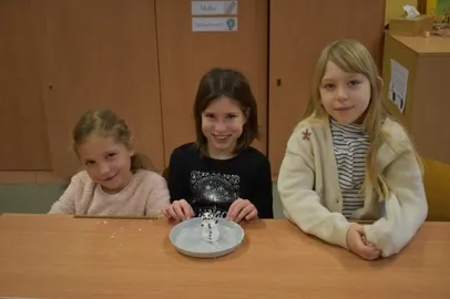 Drei junge Mädchen sitzen an einem Tisch mit einem Teller, auf dem ein Schneemann steht. Sie lächeln und posieren für ein Foto. Dahinter befinden sich Holzschränke und ein Schild an der Wand.