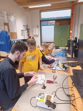 Mehrere Schüler arbeiten in einem Klassenzimmer an einem Projekt, mit einem Mann und drei Kindern. Sie haben elektronische Geräte und Werkzeuge auf dem Schreibtisch.