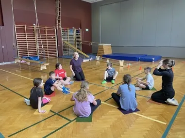 In einer Turnhalle sitzen mehrere Kinder im Kreis auf dem Boden, und zwei Frauen stehen vor ihnen. Die Frauen scheinen eine Yogastunde zu leiten. Verschiedene Übungsgeräte sind umher verstreut.