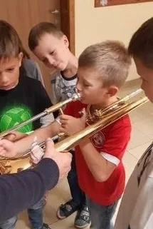 Vier Jungen stehen um einen Posaune herum. Ein Junge in Rot hält die Posaune, ein anderer in Schwarz hilft. Sie sind wahrscheinlich in einer Musikstunde.