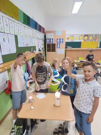Vier Jungen stehen in einem Klassenzimmer, halten Papierbasteleien und stehen um einen Tisch mit Kerzen. Das Klassenzimmer hat farbenfrohe Wände und verschiedene Lehrposter.